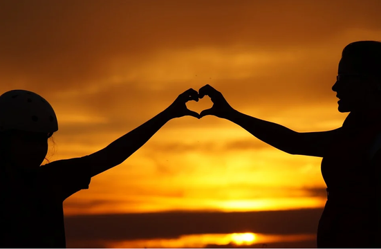 A child and adult forming a heart shape with their hands against a sunset backdrop.